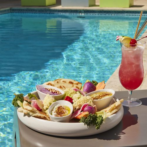 A mezze platter featuring hummus, dips, pita bread, and assorted fresh vegetables like cauliflower, radishes, and broccoli, served next to a tall, pink tropical cocktail poolside on a sunny day.