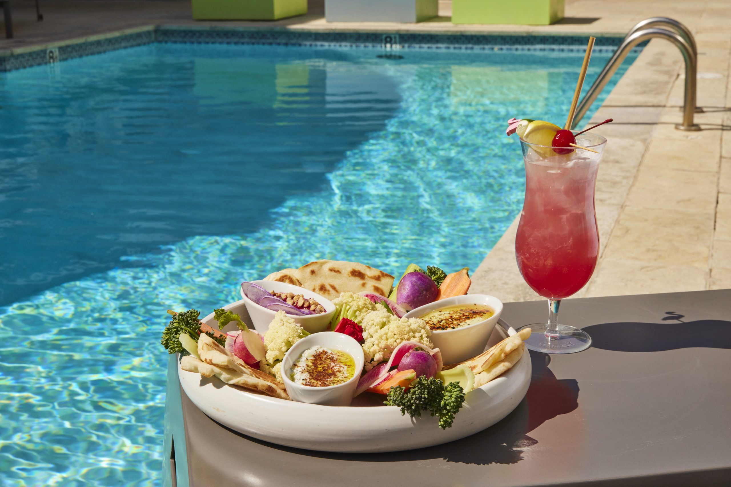 A mezze platter featuring hummus, dips, pita bread, and assorted fresh vegetables like cauliflower, radishes, and broccoli, served next to a tall, pink tropical cocktail poolside on a sunny day.