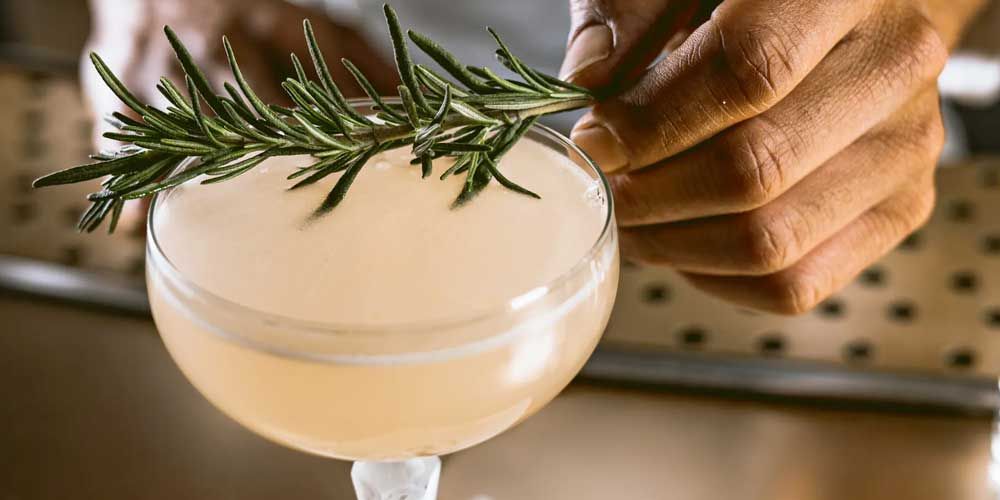 Employees Only - A faintly peach colored cocktail being adorned by a bar man with a fresh sprig of rosemary. 
