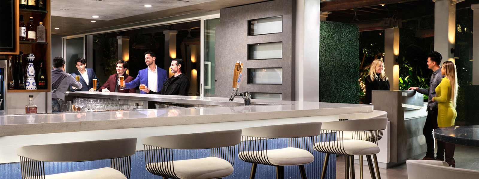 Lively modern indoor-outdoor bar with guests socializing over drinks at the counter, plush wire-frame barstools in the foreground, and a host greeting patrons near a outdoor garden entrance.