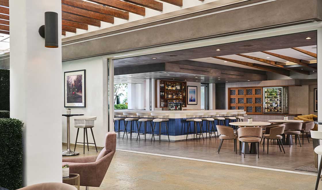 Open-air hotel bar and lounge featuring exposed wood-beamed ceilings, a central blue-accented bar with barstools, and modern dining tables leading out to a patio seating area.