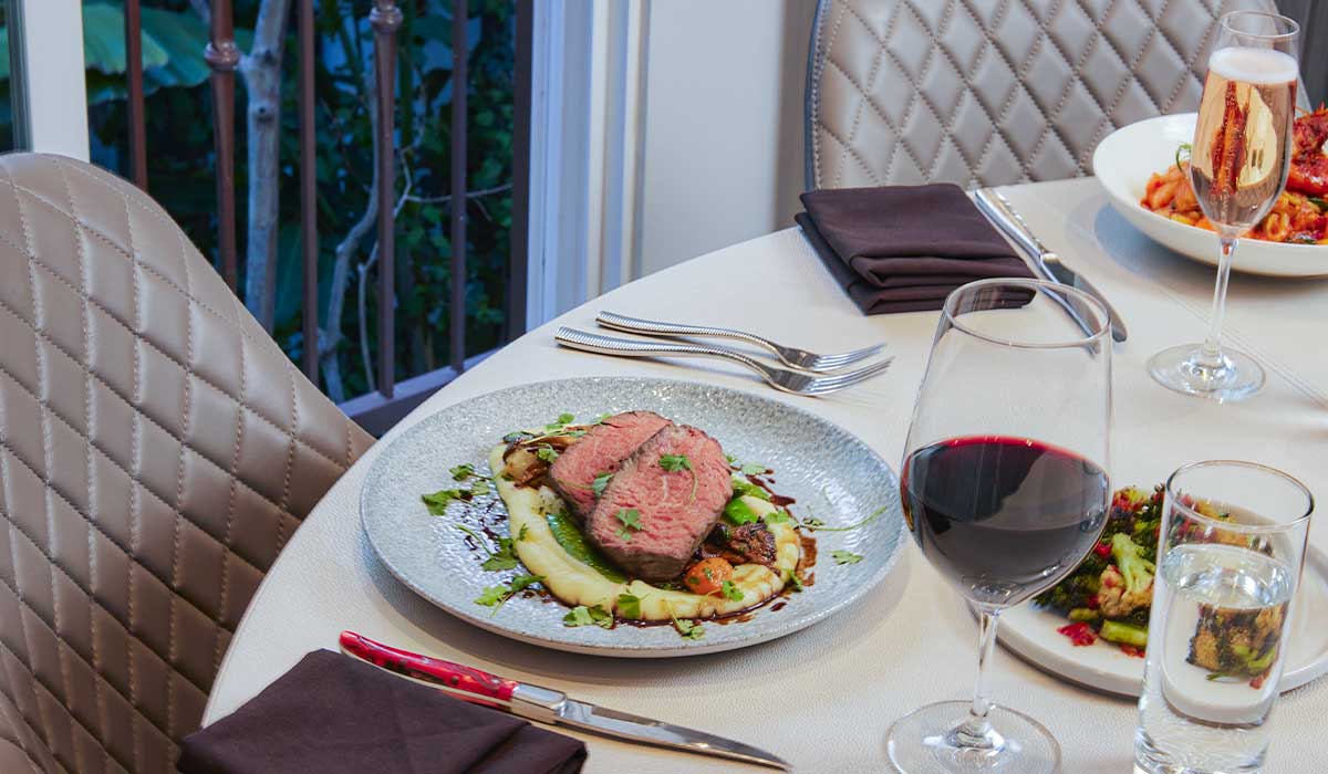 Elegantly set dinner table featuring a plated medium-rare sliced steak served over potato puree, accompanied by a glass of red wine, sparkling wine, and side dishes.