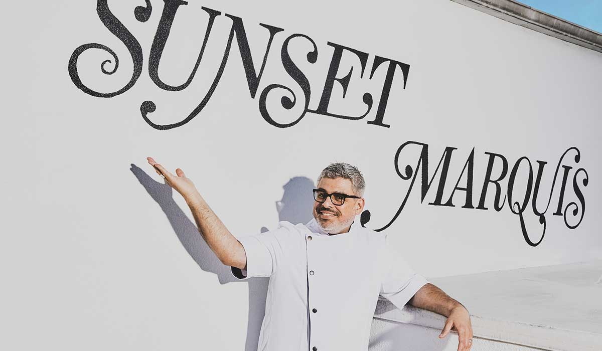 Executive Chef Luis Morales smiling and gesturing toward large 'Sunset Marquis' wall lettering outdoor in white chef attire.