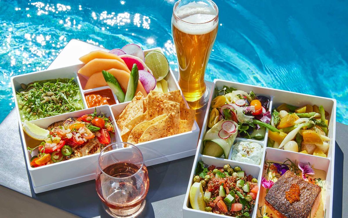 Two bento-style compartmentalized trays filled with fresh poolside dishes—including grilled chicken pico de gallo, chips and guacamole, fresh fruit, quinoa salad, and seared salmon—served alongside a tall draft beer and a glass of rosé next to a bright blue swimming pool.