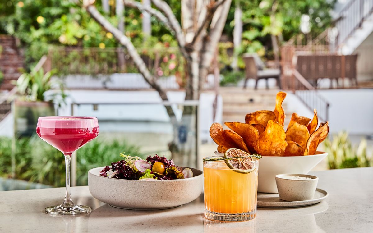 Vibrant patio dining arrangement featuring a deep pink coupe cocktail, a fresh garden salad, an orange citrus cocktail garnished with a dried lime slice, and house-made potato chips with dip.