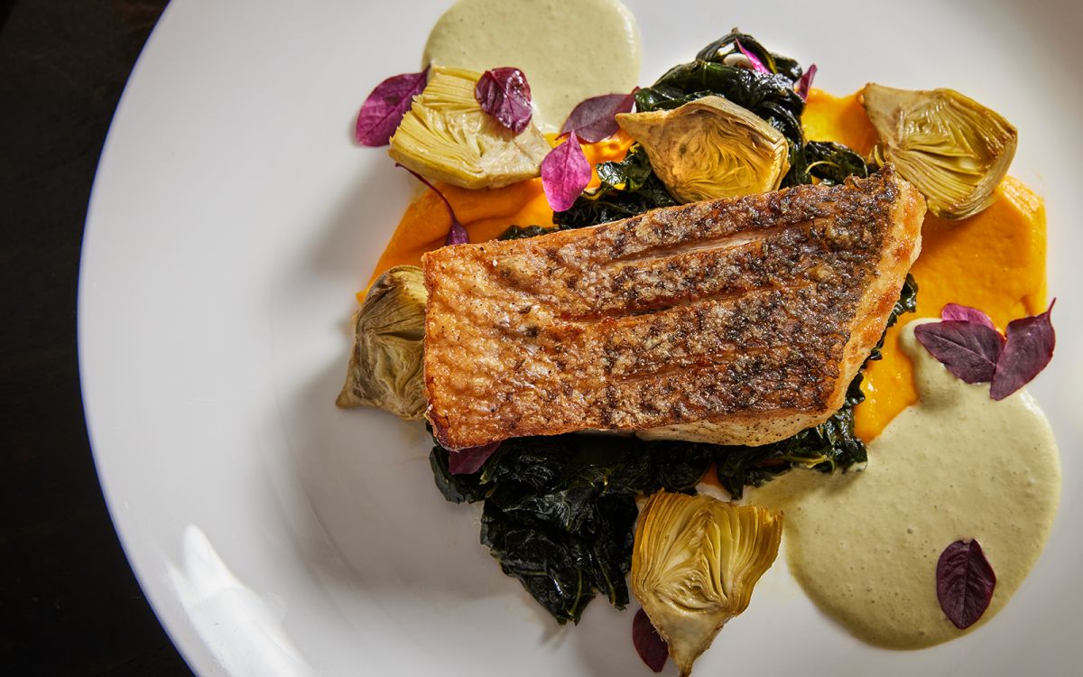 Crispy pan-seared white fish fillet served over wilted greens and orange puree, garnished with roasted artichoke hearts, purple microgreens, and a drizzle of creamy sauce.