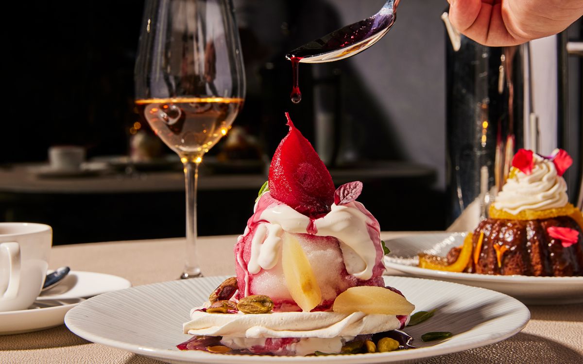 Berry syrup being drizzled from a spoon onto a poached pear pavlova dessert with whipped cream and pistachios, served alongside dessert wine and coffee.