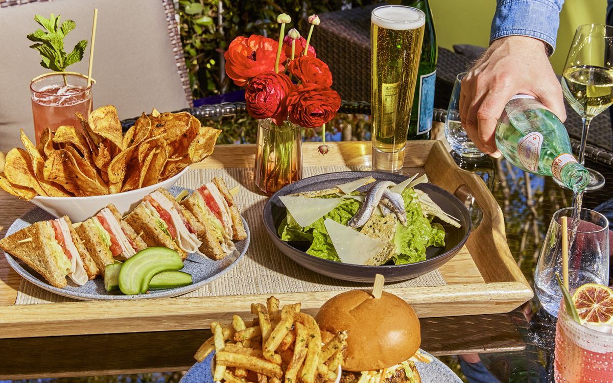 Outdoor patio dining tray featuring club sandwiches with house-made potato chips, a Caesar salad topped with anchovies and parmesan, a burger with french fries, craft drinks, and sparkling water being poured into a glass.