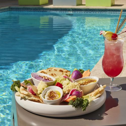 A mezze platter featuring hummus, dips, pita bread, and assorted fresh vegetables like cauliflower, radishes, and broccoli, served next to a tall, pink tropical cocktail poolside on a sunny day.