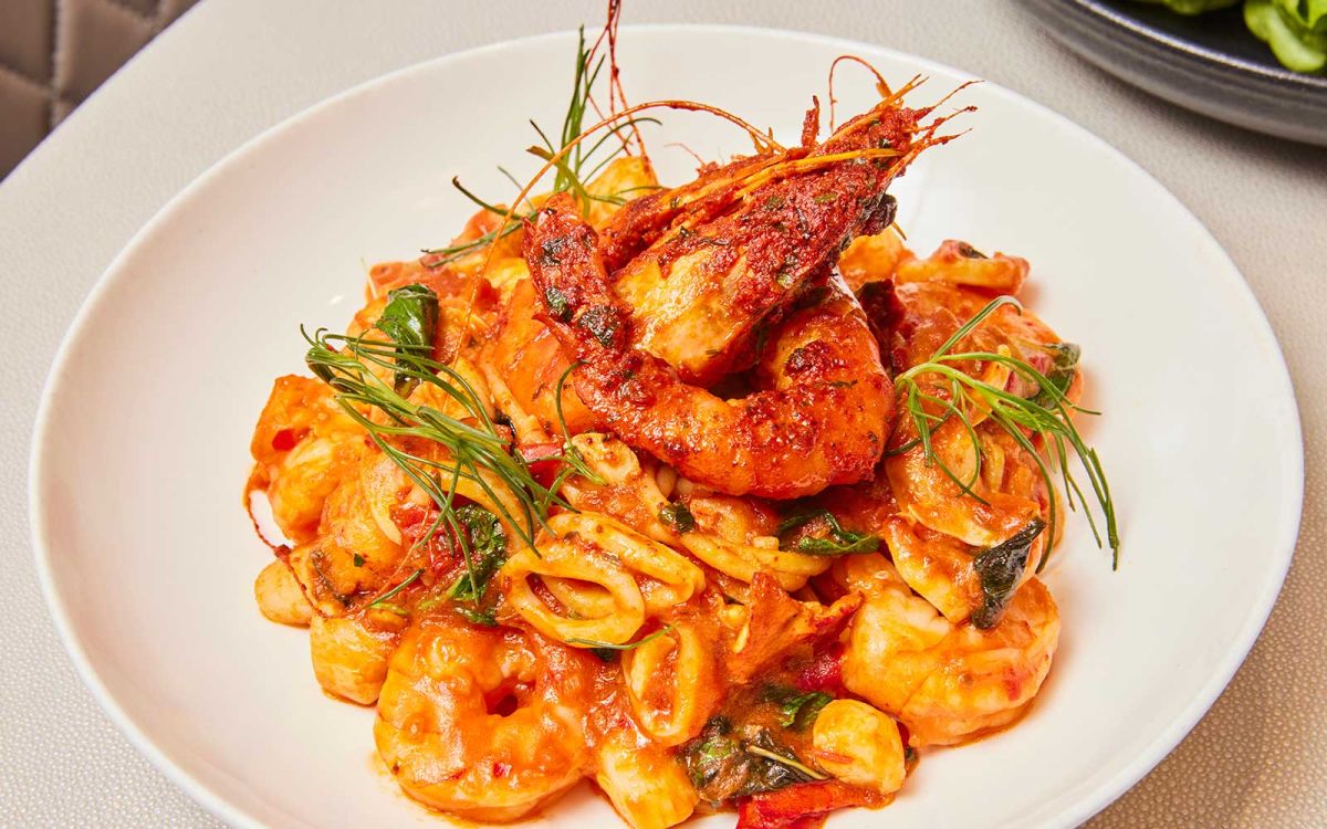 Elegantly plated seafood pasta dish in a rich tomato sauce, topped with jumbo prawns, calamari rings, fresh herbs, and microgreens in a shallow white bowl.