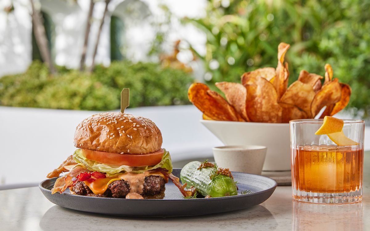 Gourmet bacon cheeseburger on a sesame seed bun served with a side pickle, house-made potato chips, and an Old Fashioned cocktail on an outdoor patio.