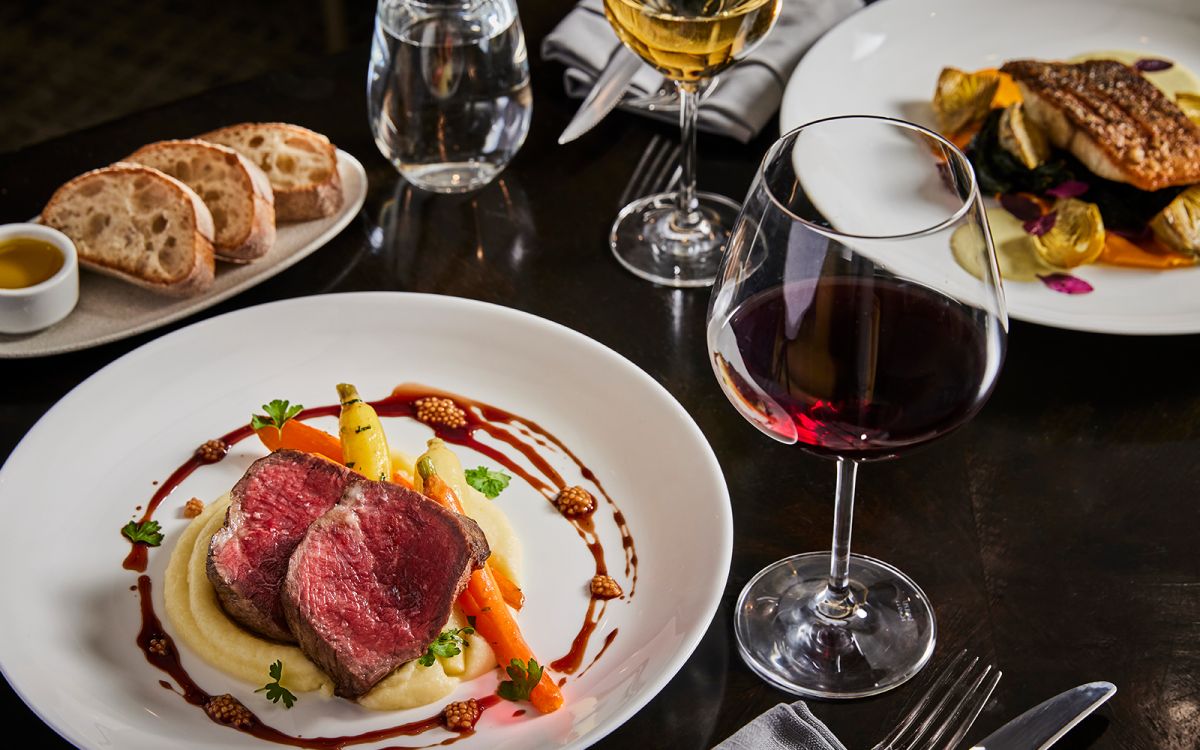 Fine dining dinner setting featuring sliced medium-rare steak over potato puree with glazed baby carrots and red wine sauce, served alongside a glass of red wine, sliced crusty bread, olive oil, and a pan-seared fish entree.