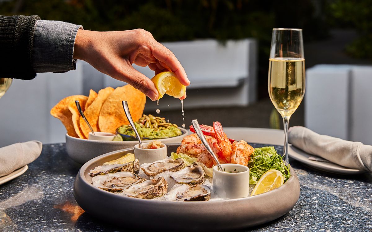 Hand squeezing fresh lemon juice over a raw bar platter featuring oysters on the half shell and jumbo shrimp cocktail, served alongside guacamole with tortilla chips and a glass of champagne.