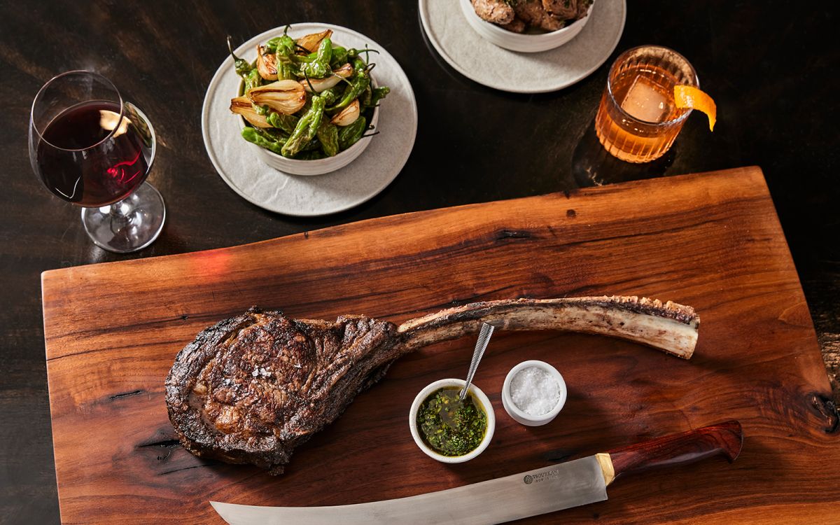 Large grilled tomahawk ribeye steak served on a wooden board with chimichurri sauce, sea salt, a carving knife, blistered shishito peppers, an Old Fashioned cocktail, and a glass of red wine.