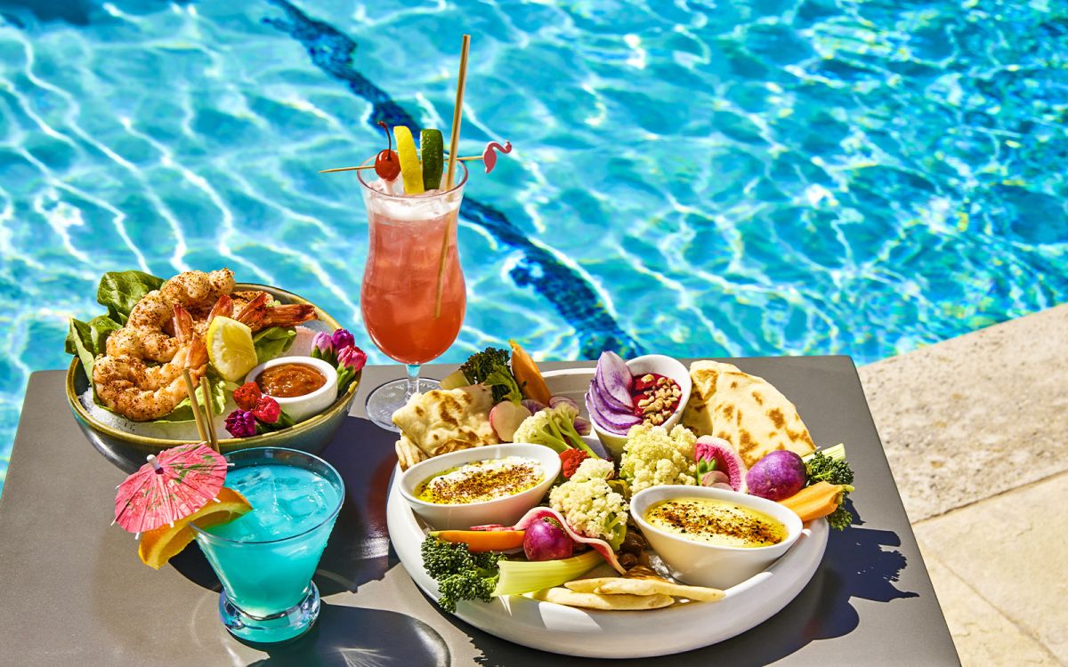 Poolside food spread featuring a Mediterranean dip platter with pita and fresh vegetables, grilled shrimp cocktail, a bright blue tropical cocktail with a paper umbrella, and a pink hurricane drink by a sunny swimming pool.