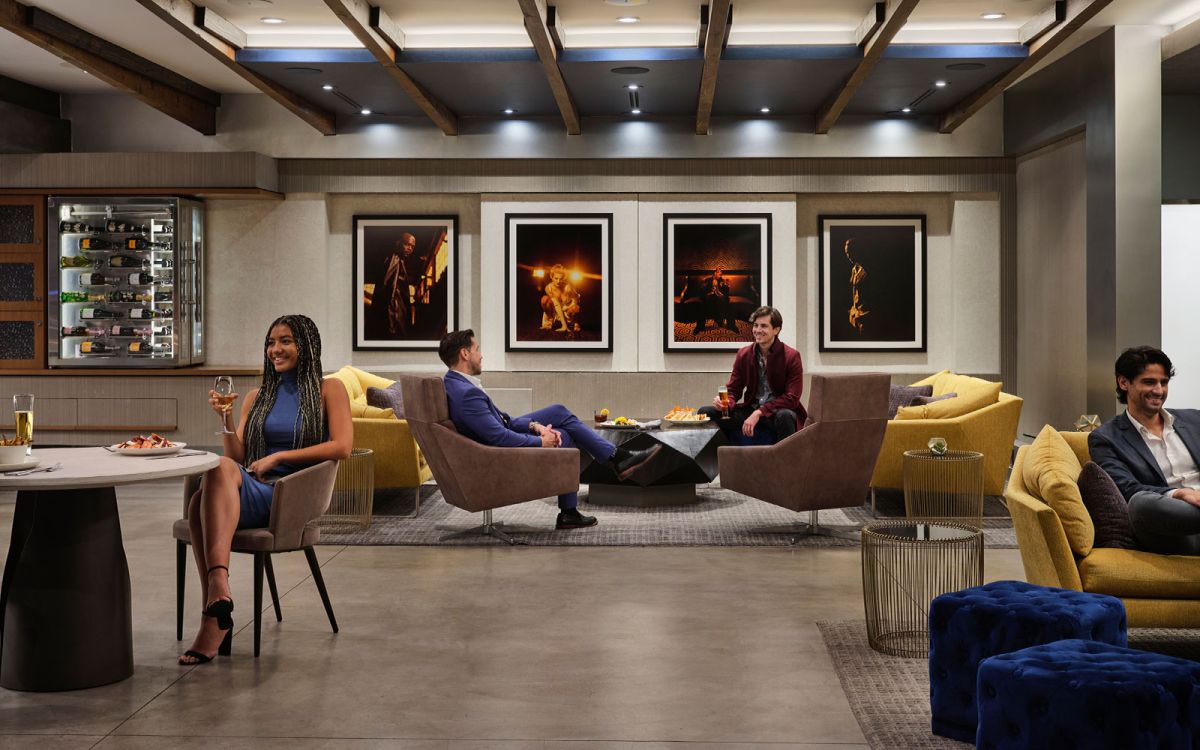 Guests relaxing and socializing in a stylish upscale lounge with plush armchairs, framed wall art, exposed wooden ceiling beams, and a modern wine display."